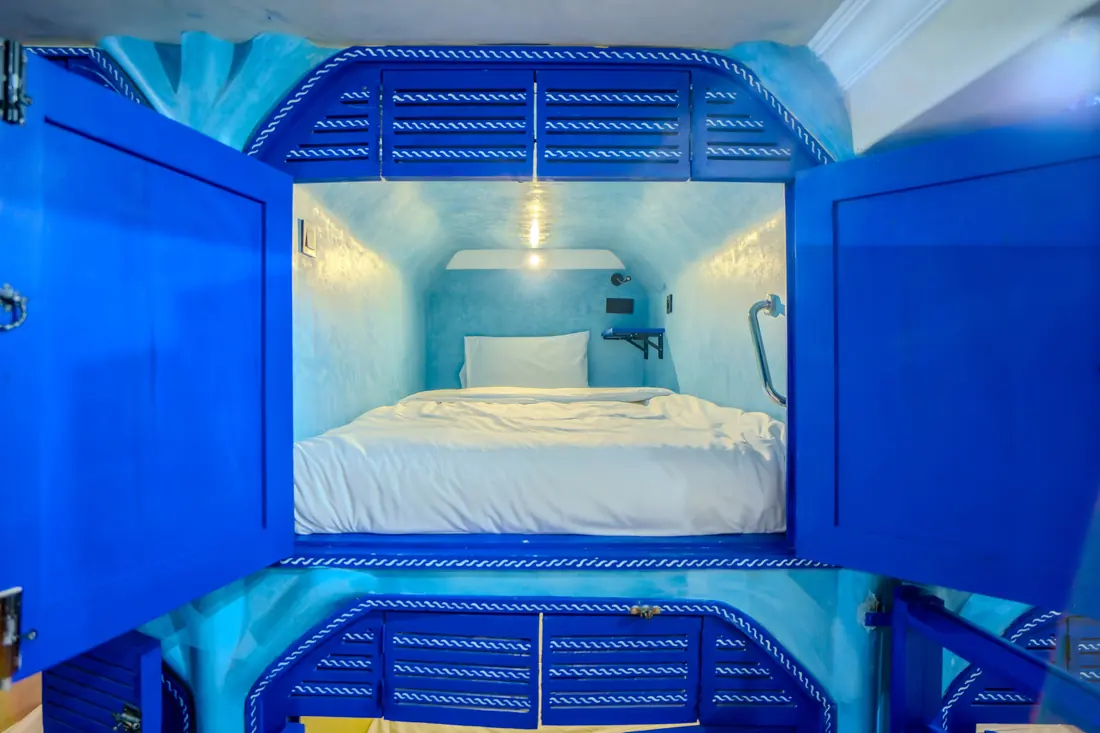 Une cabine fermée par des volets bleus, avec un lit deux places, une liseuse et une tablette