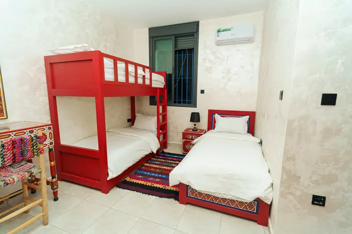 Une chambre avec un lit superposé rouge, un lit simple à tête peinte, un tapis et une fenêtre