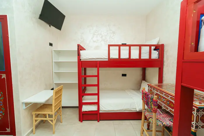 Une chambre avec un lit superposé rouge, une étagère blanche, un bureau et un téléviseur mural