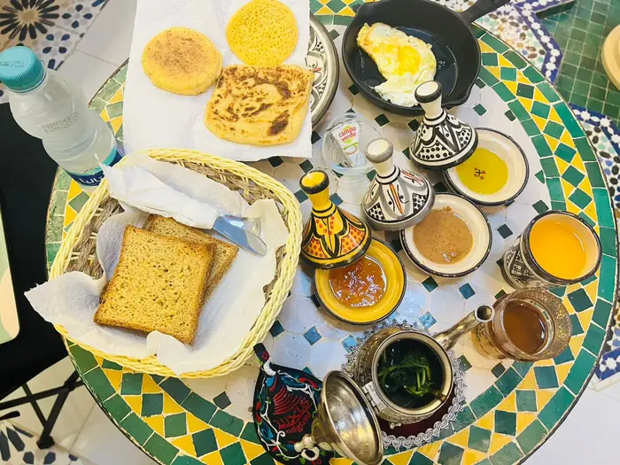 Une table de petit-déjeuner en zellige : beghrir, msemen, harcha, œuf au plat, huile d'olive, confiture, jus d'orange et thé